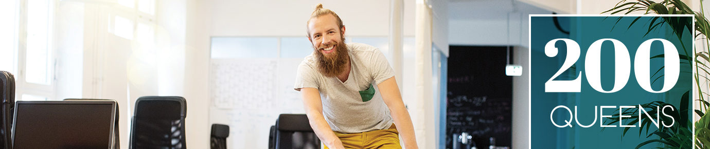 A young and bearded man wearing bright yellow pants and who is smiling while riding a bike. A large copy block on the right side that says 200 Queens