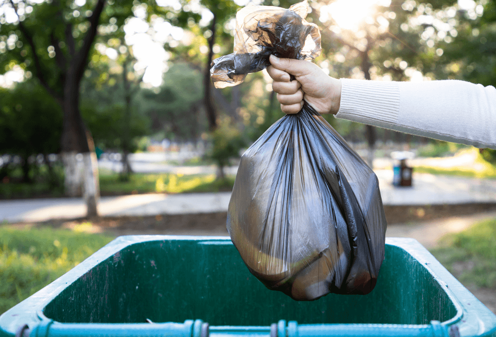 A hand holding a garbage bag over a green garbage bin